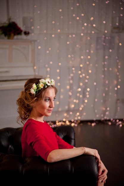 A blonde girl in a red dress and with flowers in her hair sits on a leather sofa