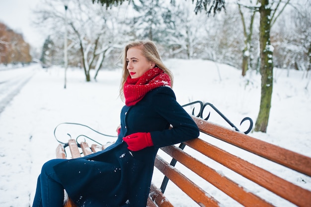 Blonde girl in red scarf and coat sitting at bench on winter day.