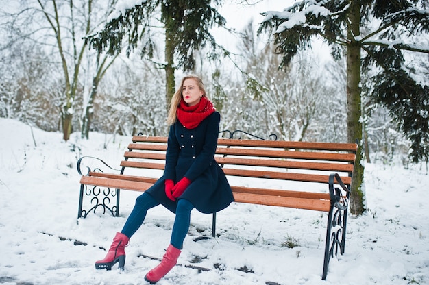 Blonde girl in red scarf and coat sitting at bench on winter day.