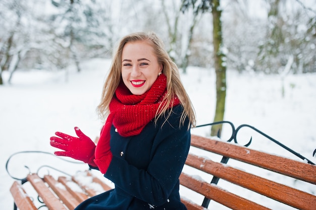 Блондинка в красном шарфе и пальто, сидя на скамейке в зимний день.