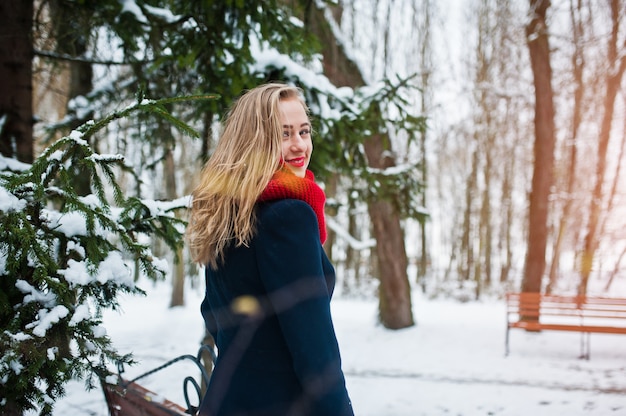 Blonde girl in red scarf and coat walking at park on winter day.