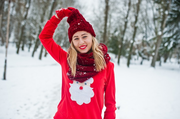 Blonde girl in red scarf, hat and santas sweater posing at park on winter day