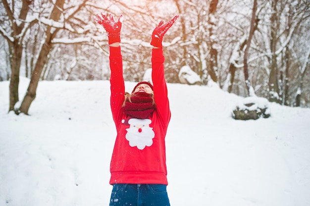 Blonde girl in red scarf, hat and santas sweater posing at park on winter day.