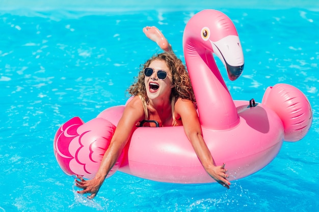 Foto ragazza bionda che riposa nella piscina estiva su un fenicottero rosa gonfiabile in costume da bagno.