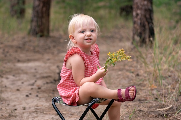 Blonde girl sitting on chair with flower in hands in forest. Happy child on the field with wildflower. Summer mood.