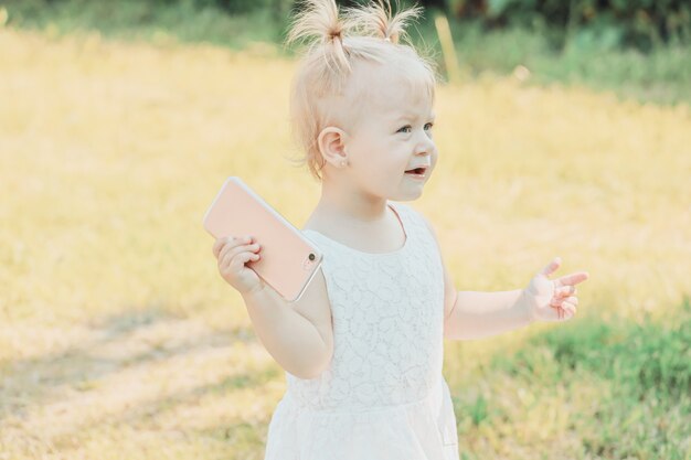 Blonde girl standing on the grass in the summer garden with a smartphone in her hands. High quality photo