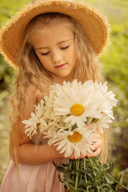 a blonde girl in a straw hat in a pink linen dress holds a bouquet of daisies in her hands