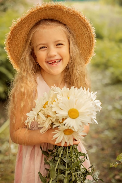 A blonde girl in a straw hat in a pink linen dress holds a bouquet of daisies in her hands