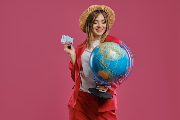 Blonde girl in straw hat white blouse and red pantsuit she is smiling holding globe passport