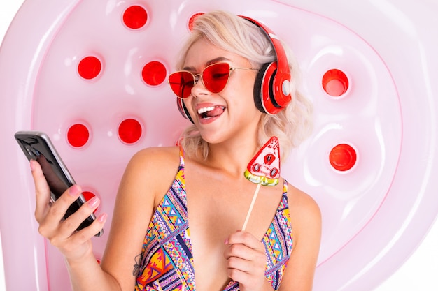 блондинка в купальнике и темных очках с леденцом и телефоном в руках на плавательном матрасе слушает музыку в наушниках