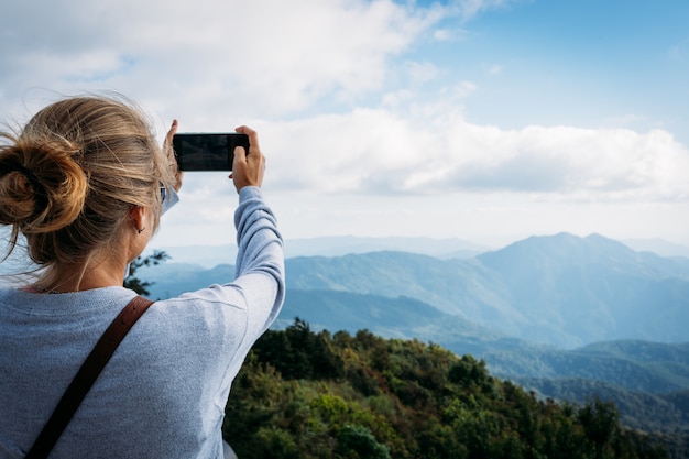 Blonde girl taking pictures with the mobile of the horizon views in the mountains