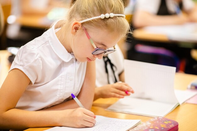 Ragazza bionda con grandi occhiali seduto in aula, studiando, sorridendo. istruzione sulla scuola elementare, primo giorno a scuola