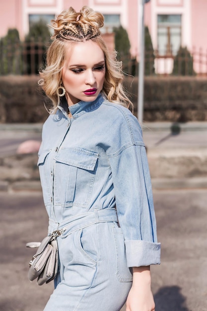 Blonde girl with bright makeup and hairstyle in denim jumpsuit posing on a city street