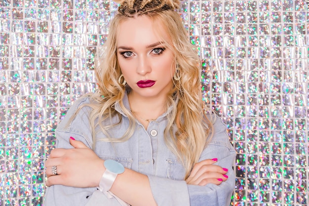 Blonde girl with bright makeup and hairstyle in denim overalls posing on a glitter background