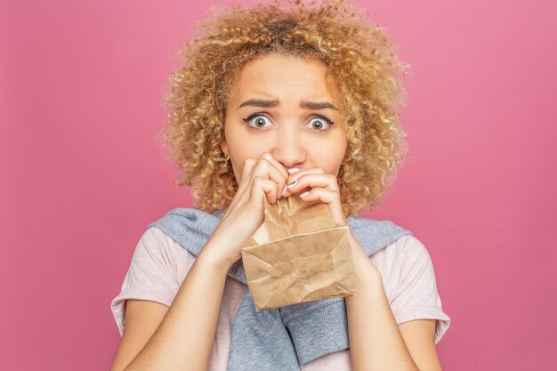 Photo blonde girl with a curly hair is looking straight and trying to blow the paper bag.