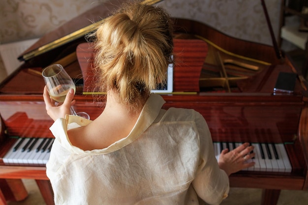 Blonde girl with glass of wine plays retro piano watches video on tablet