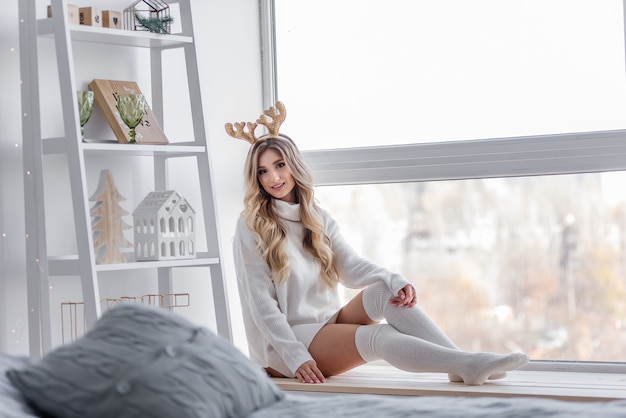 A blonde girl with golden Christmas deer horns in a gray knitted sweater, long leggings sits on the windowsill by the panoramic window. There is a white rack with home figurines. Copy space blurred