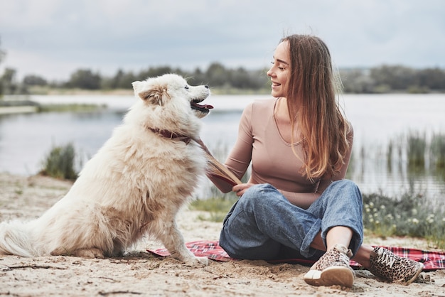 彼女のかわいい白い犬と一緒のブロンドの女の子は、ビーチで過ごすのに素晴らしい時間を過ごします。