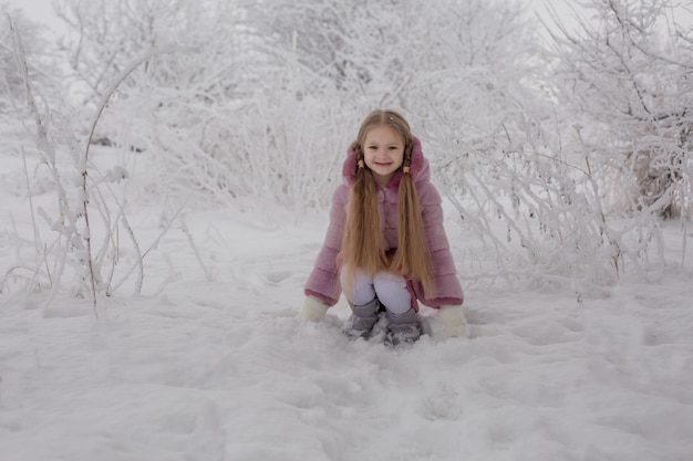 ピンクの毛皮のコートを着た長い髪のブロンドの女の子が雪に覆われた冬の公園に座っています