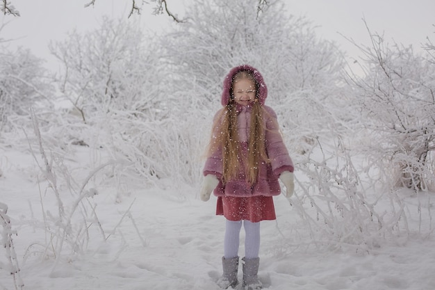 雪に覆われた冬の公園でピンクの毛皮のコートを着た長い髪のブロンドの女の子