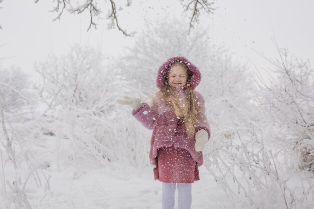 雪に覆われた冬の公園でピンクの毛皮のコートを着た長い髪のブロンドの女の子