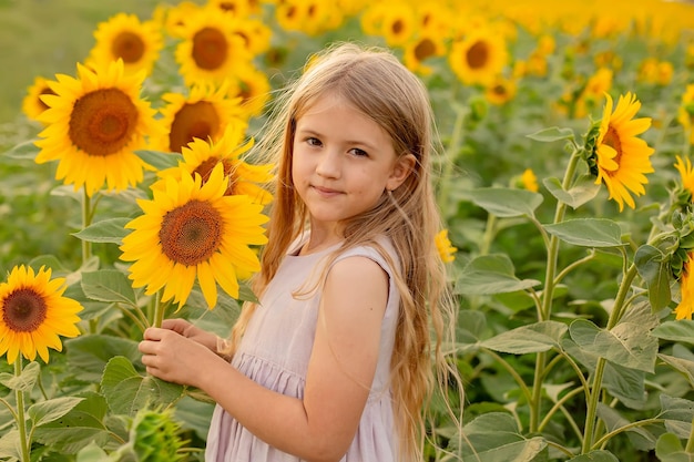 a blonde girl with long hair in a pink linen dress stands in a field with sunflowers