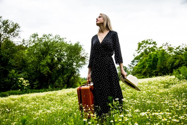 Blonde girl with suitcase and hat in chamomiles flowers field in summer time