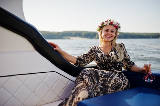 Blonde girl in wreath sitting on yacht at hen party with champagne.