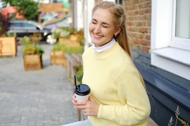 A blonde girl in a yellow sweater and jeans sits on a bench with a cup of takeaway coffee the girl l