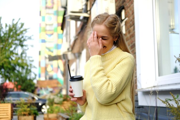 A blonde girl in a yellow sweater and jeans sits on a bench with a cup of takeaway coffee the girl l