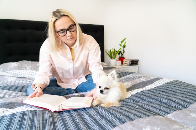 ブロンドの髪の女性は犬と一緒に居心地の良いベッドの上に座って、開いた本を持って読んでいます。横になっている女性が自宅のソファーでリラックス。レジャーと快適な人々の概念。隔離するための時間。