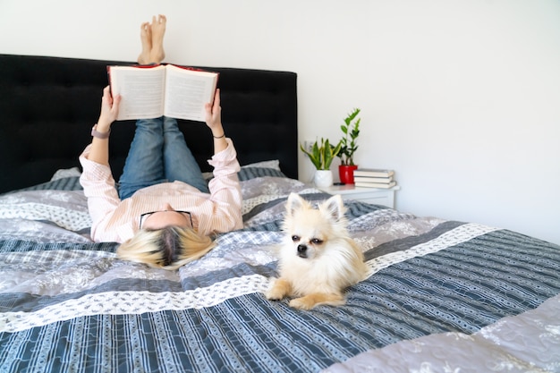 Светлые волосы женщина сидит на уютной кровати с собакой, держа открытую книгу и чтение. Лежа женщина расслабляющий на диване у себя дома. Концепция отдыха и комфорта людей. Время для себя в карантине.