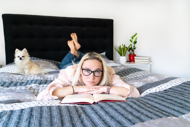 Blonde hair woman sitting on cozy bed with dog, holding open book and reading. Lying woman relaxing on sofa at home. leisure and comfort people concept. Time for yourself in quarantine.