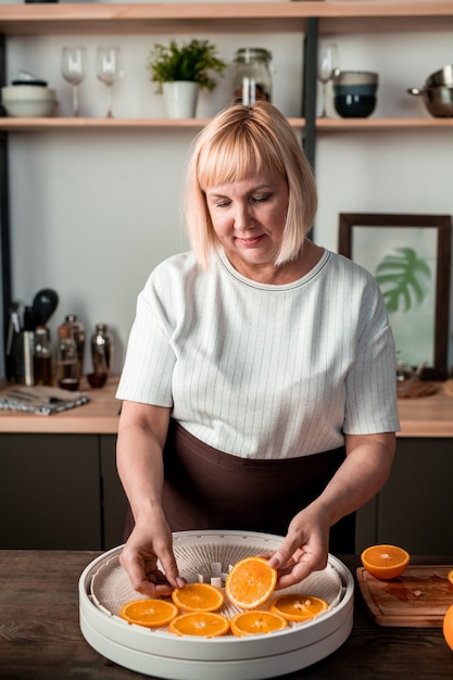 Blonde huisvrouw van middelbare leeftijd in t-shirt en schort die naar je kijkt terwijl ze bij de keukentafel staat en sinaasappelschijfjes op de fruitdroger legt