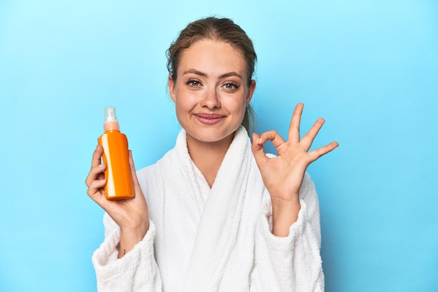 Blonde in badjas met zonnebrandcrème in studio, vrolijk en zelfverzekerd met ok gebaar