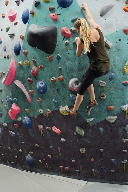 Blonde jonge actieve vrouwelijke bedrijf door kunstmatige rotsen op de muur tijdens klimoefening in de sportschool of extreme sporten centrum