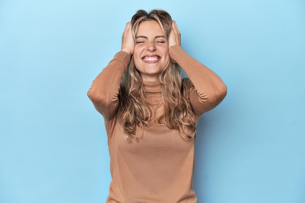 Blonde jonge blanke vrouw in blauwe studio lacht vreugdevol met handen op het hoofd Geluk concept