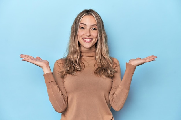 Blonde jonge blanke vrouw in blauwe studio maakt schaal met armen voelt zich gelukkig en zelfverzekerd