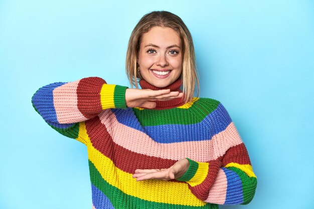 Blonde jonge vrouw in een veelkleurig sweatshirt op blauwe achtergrond die iets met beide handen vasthoudt