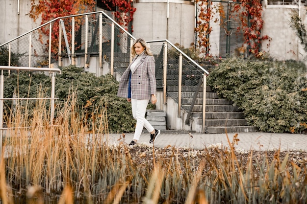 Blonde jonge vrouw in lichte trui, witte spijkerbroek en grijze jas die op afstand buiten loopt, mogelijk in de stad of stad in de vroege herfst met zijn gele gras, groene en roodachtige bladeren