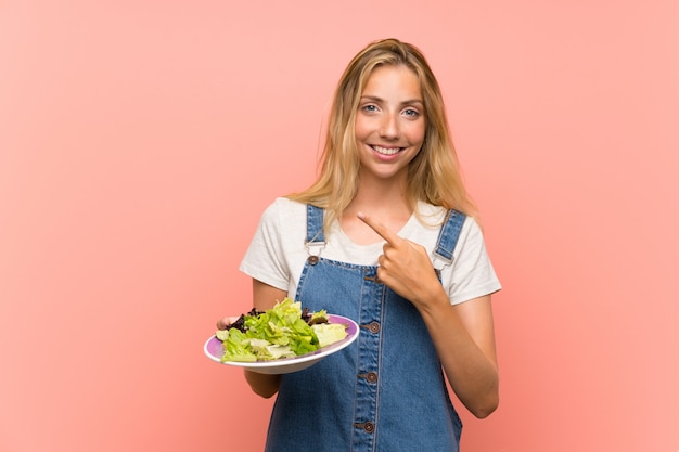 Blonde jonge vrouw met salade over geïsoleerde roze muur die aan de kant richten om een product te presenteren