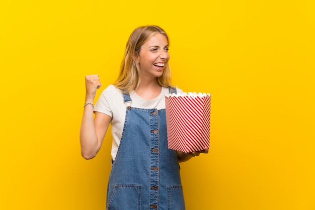 Blonde jonge vrouw over geïsoleerde gele muur die een kom popcorns houden