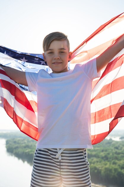 Blonde jongen die de nationale vlag van de VS buiten zwaait over de blauwe lucht aan de rivieroever