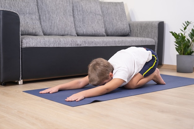 Blonde jongen gaat sporten op yogamat. Lichamelijke activiteit van het kind. Sport gezonde levensstijl actieve vrije tijd thuis tijdens COVID-19 quarantaine.