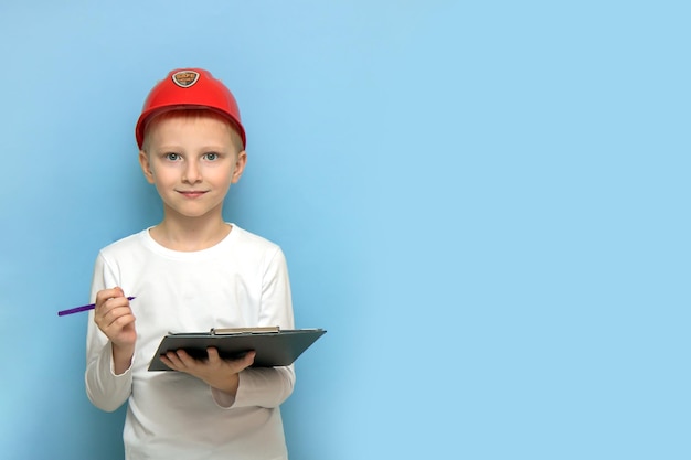 Blonde jongen in een beschermende bouwhelm met een tablet en een pen op een blauwe achtergrond met kopieerruimte die op een bouwplaats controleert