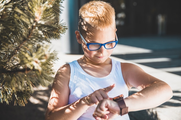 Foto blonde jongen in glazen kijkt op zijn horloge in het park.