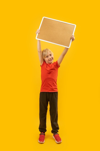 Blonde jongen in rood T-shirt houdt leeg kurkbord in handen boven zijn hoofd Portret van kind met blanco vel Kopieer ruimte Verticaal frame