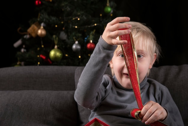 Blonde jongen met kerstklatergoud in zijn handen zittend op de bank tegen de achtergrond van de kerstboom.