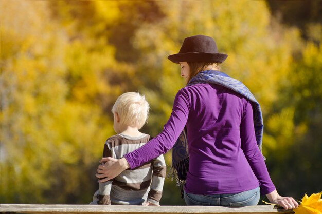 Blonde jongen met zijn moeder in de hoed zittend op de bank. Herfst bos. Achteraanzicht