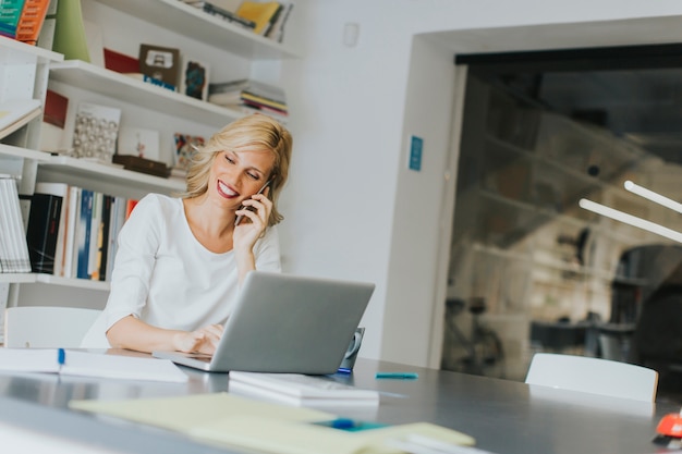 Blonde Kaukasische onderneemster die haar mobiele telefoon met behulp van en bij haar bureau zit